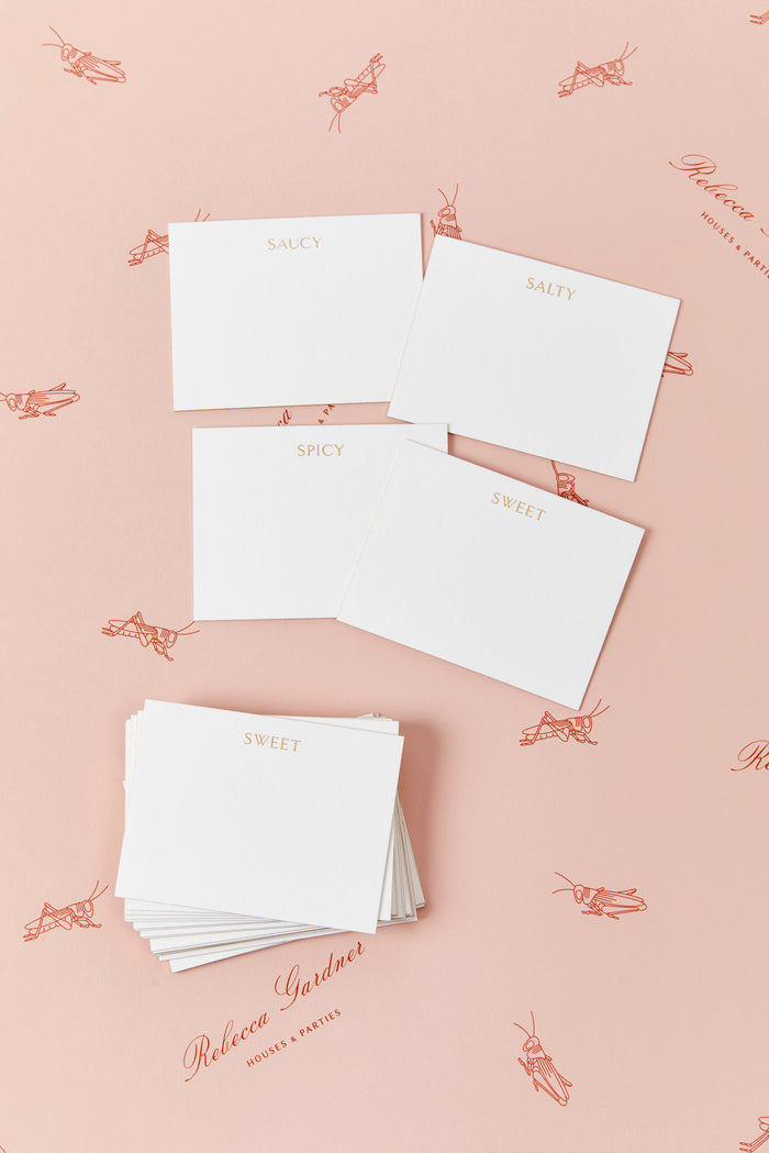 Set of white place cards displayed on top of a light pink background. Place cards are white with gold engraved lettering across the top featuring a variety of words including "saucy", "salty", "spicy", and "sweet." Four cards with each word are displayed next to a stack of several cards.