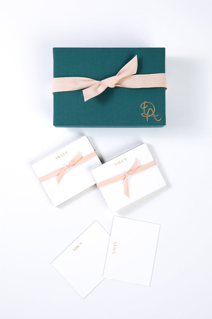 Box and set of white place cards displayed on a white background. Place cards are white with gold engraved lettering across the top featuring a variety of words including "saucy", "salty", "spicy", and "sweet." Four cards with each word are displayed next to a blue box with a gold logo and light pink ribbon.